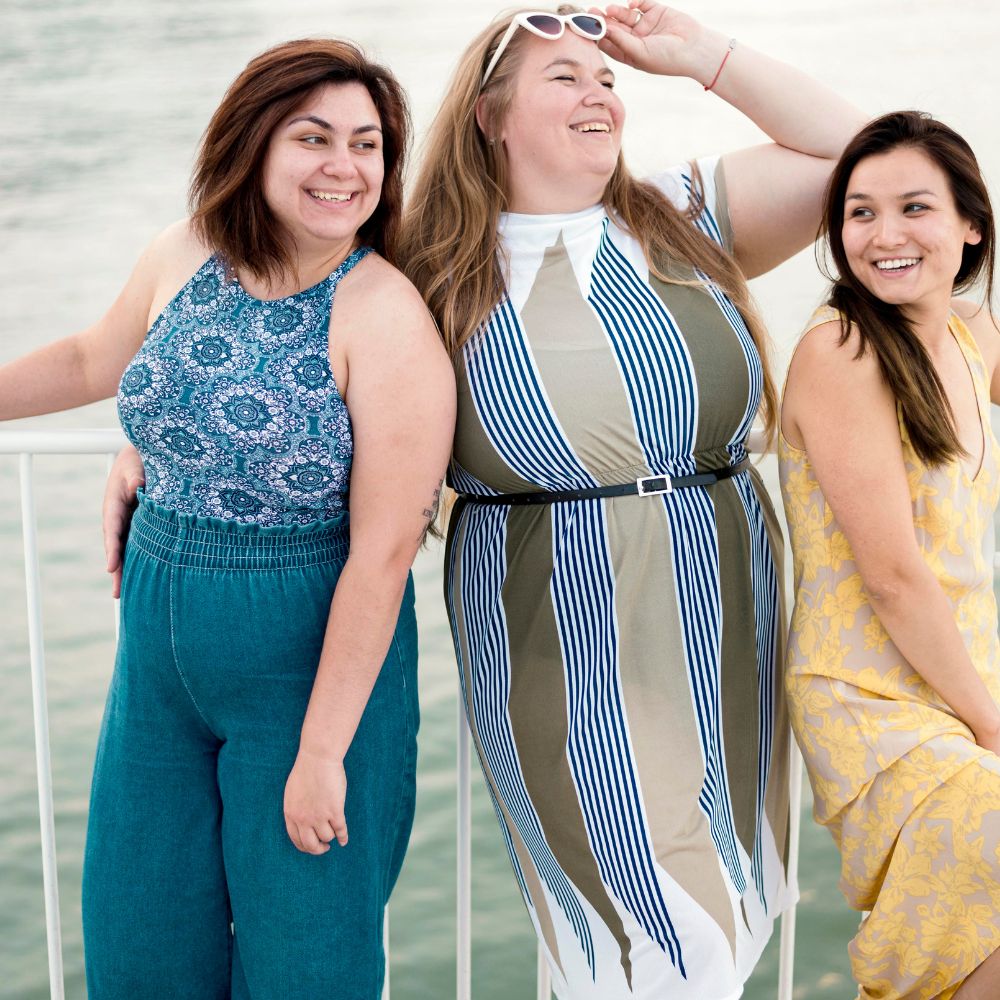 Three women posing together by a body of water, wearing stylish outfits.