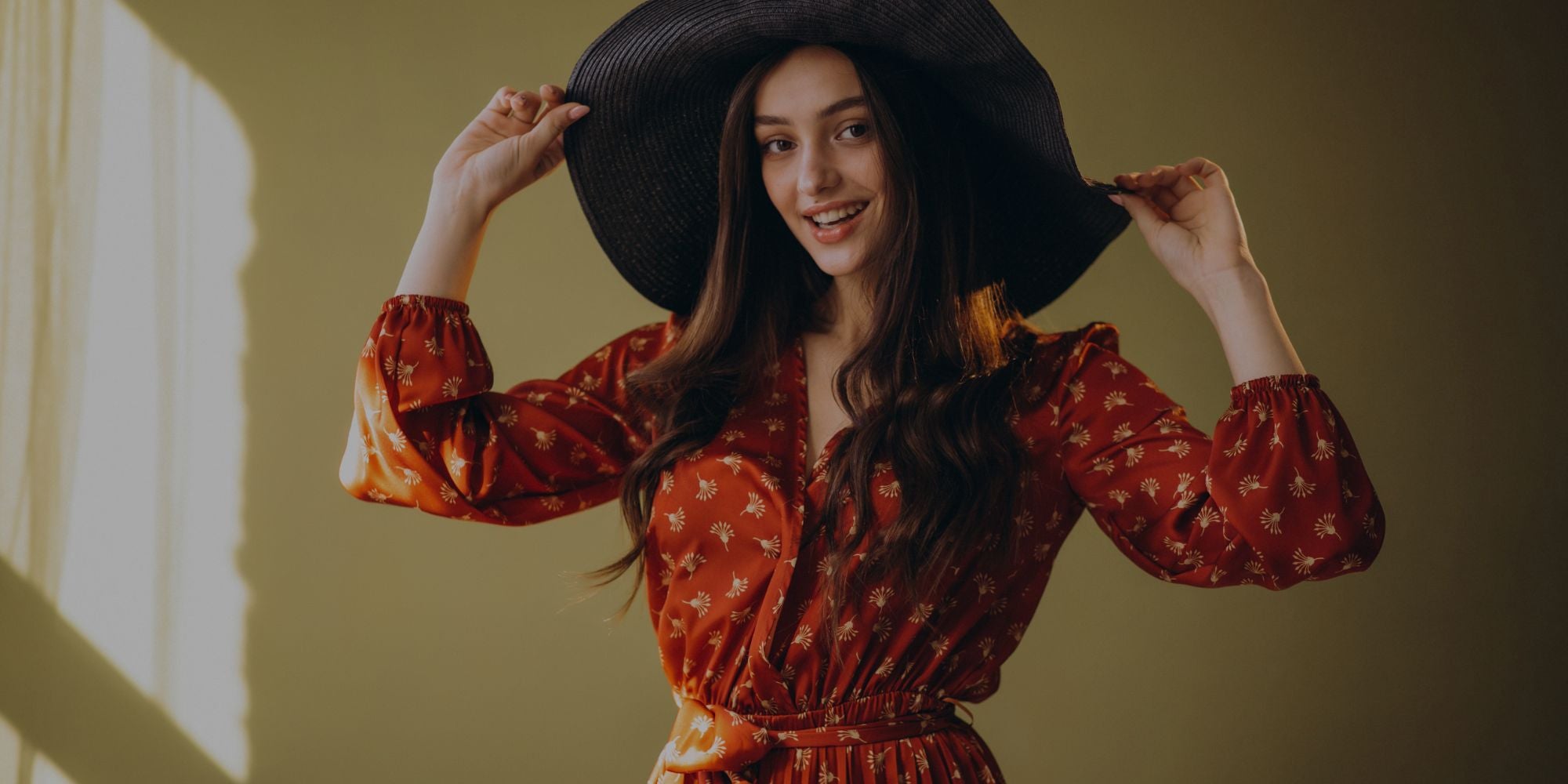 Woman in a red dress holding a black hat indoors with a warm light source.